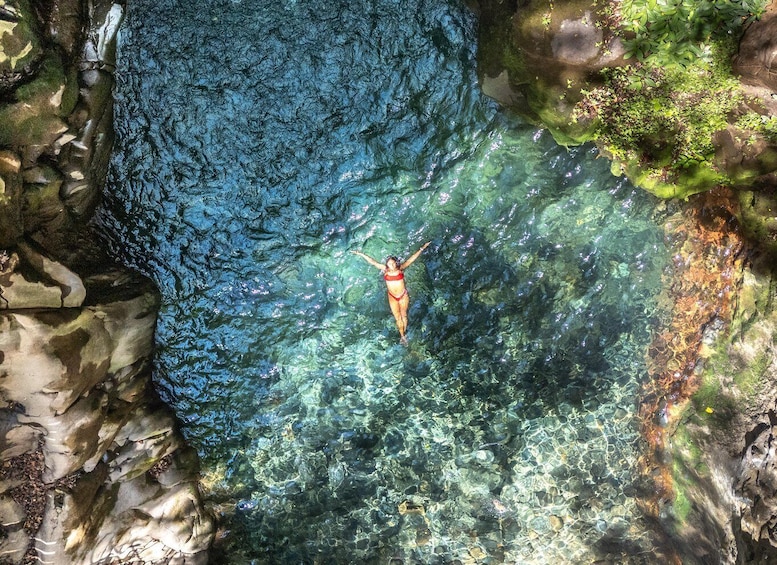 A person swimming in a clear blue pool surrounded by rocks and greenery. 1