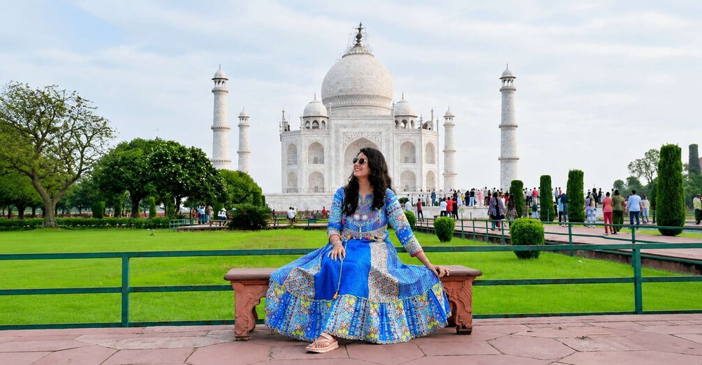 A woman in a blue dress sitting on a bench with the Taj Mahal in the background. 1