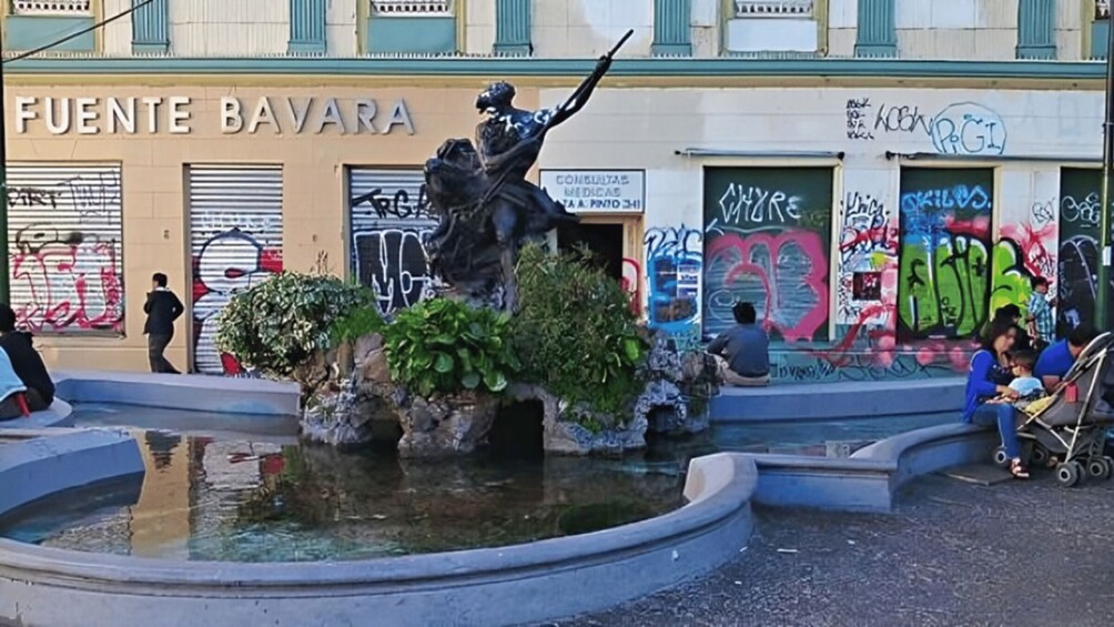 A statue in a fountain, a building with 'FUENTE BAVARA' signage, and graffiti on the walls. 1