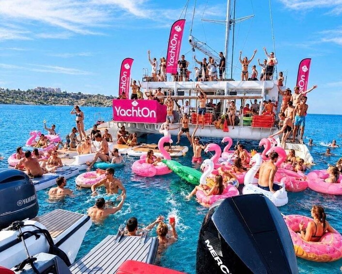 A yacht party with people on inflatable flamingos in the water, a yacht with 'YachtOn' flags, and a clear blue sky. 1