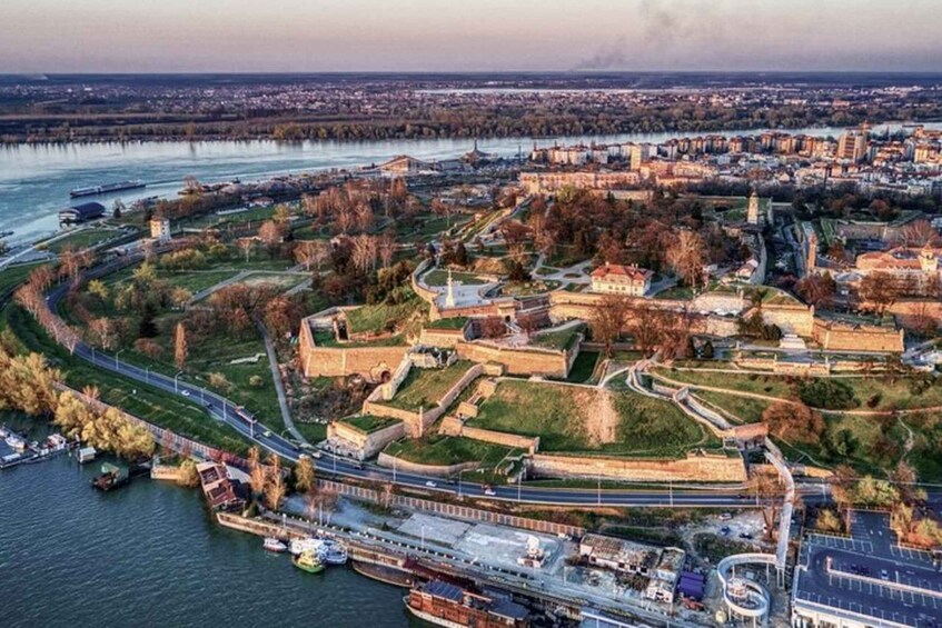 An aerial view of a historic fortification complex by a river, surrounded by urban development and modern infrastructure. 1