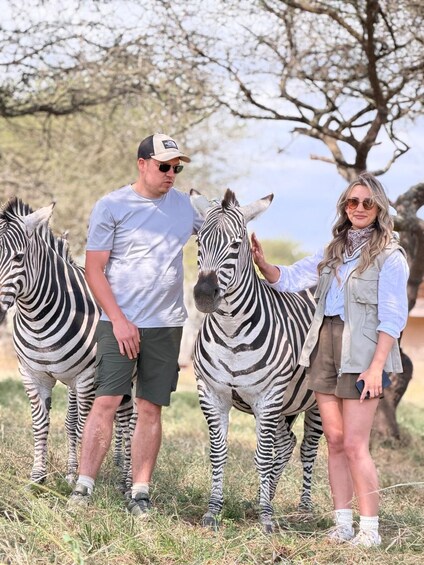 Two zebras and two people standing in a grassy field with trees in the background. 5