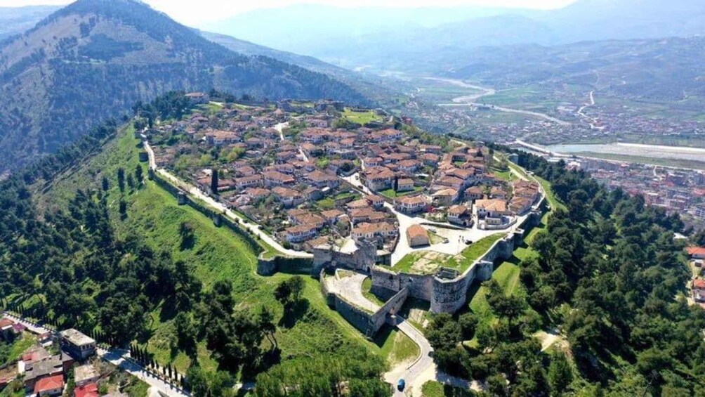 Vista aerea di una città storica cinta da mura, con una fortezza centrale, circondata da rigogliosa vegetazione e un paesaggio montuoso. 4