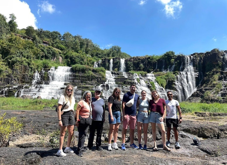 A group of people standing in front of a multi-tiered waterfall with lush greenery in the background. 1
