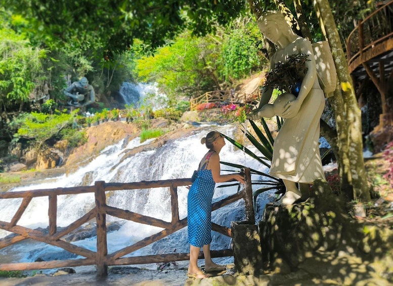 A woman in a polka dot dress standing by a waterfall, with a statue and lush greenery in the background. 2