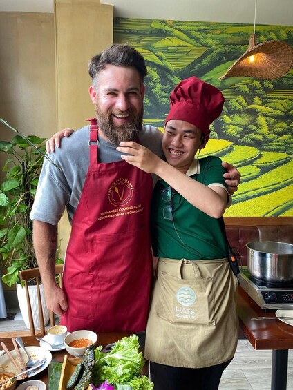 Two people in a kitchen, one wearing a red apron and the other a green shirt, with a painting of a landscape in the background. 2