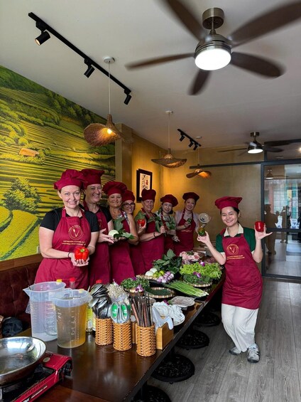 A group of chefs in red aprons and hats holding vegetables in a kitchen with a mural and various cooking utensils on the counter. 1