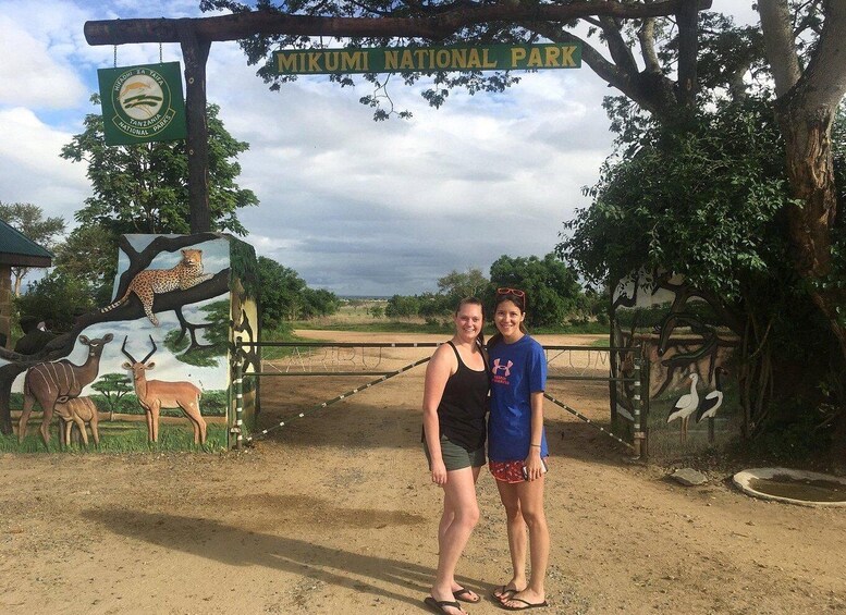 Two people standing in front of a sign that reads "Mikumi National Park" with a leopard and other wildlife illustrations. 1