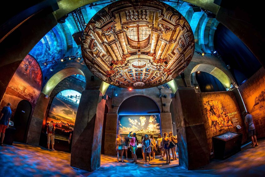 A large, ornate ceiling with intricate designs and a central circular element. People are gathered in front of a large digital display showing a sunset scene. 4