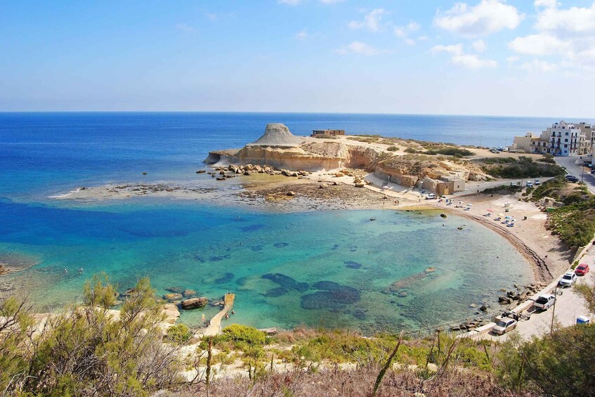 A coastal landscape with clear blue waters, a sandy beach, and a rocky outcrop with buildings. 1