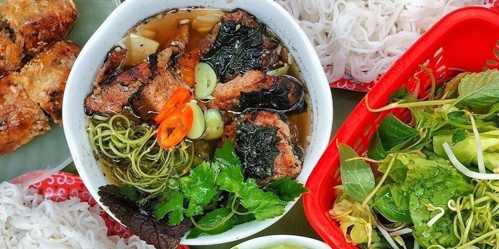 A bowl of soup with noodles, meat, and vegetables, surrounded by rice and fresh herbs. 1