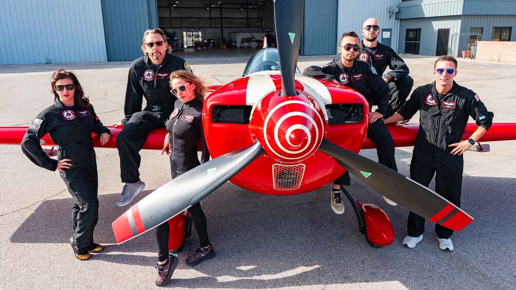 A group of people in flight suits posing with a red and white propeller plane. 4