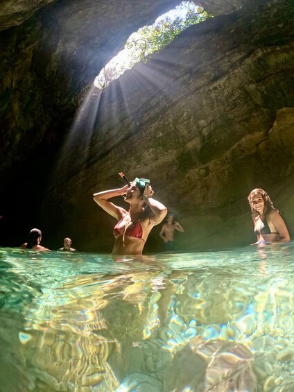 A group of people swimming in a natural pool with sunlight streaming through an opening above. 2