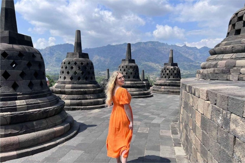 A person in an orange dress walking between stone structures with a mountainous backdrop. 3