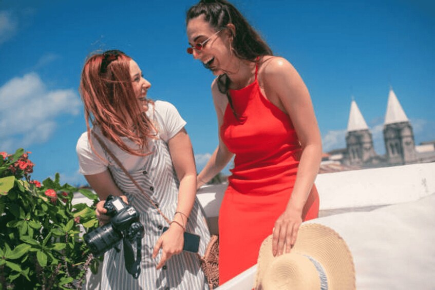 Two women, one in a red dress and the other in a striped dress, on a rooftop with a camera and a straw hat. 3