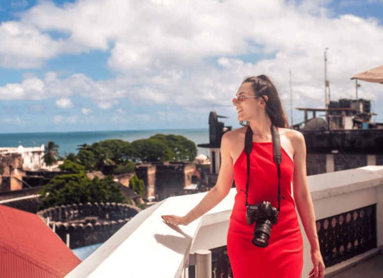 A person in a red dress standing on a balcony overlooking a coastal cityscape with buildings and a clear sky. 2