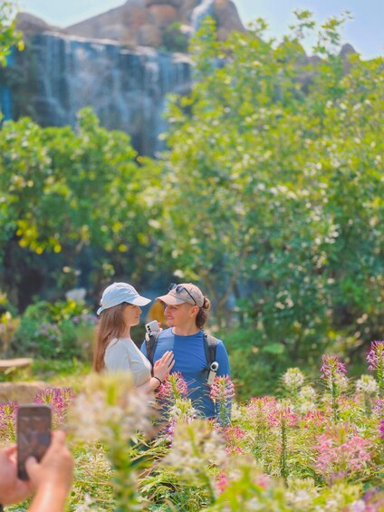Two people in a garden with flowering plants and a waterfall in the background. 1