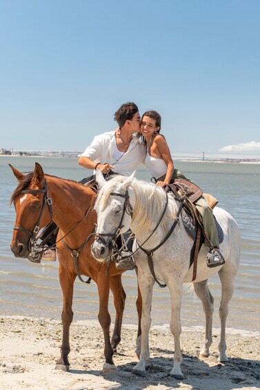 Two horses, one brown and one white, with riders on a beach near water. 3