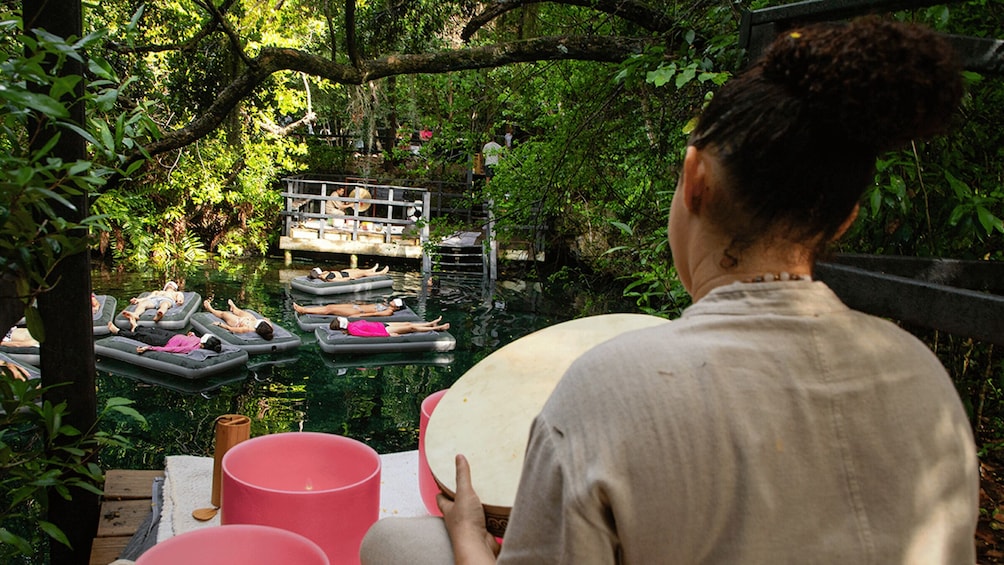 A person holding a large round object, possibly a drum, overlooking a pond with floating platforms where people are lying down. 5