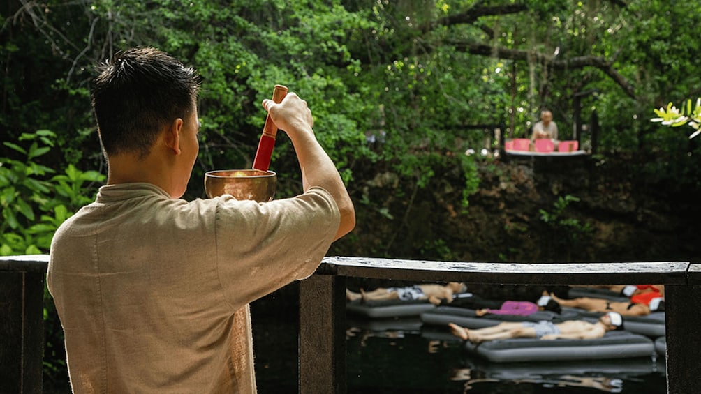 A man holding a drink and a camera, overlooking a group of people floating on water in a forested area. 1