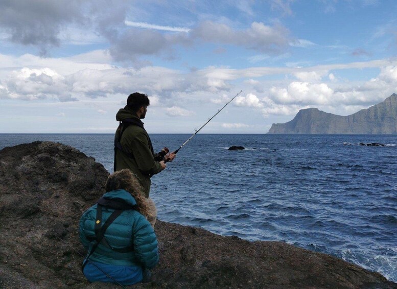 Two people fishing by the sea with a rocky shore and a mountain in the background. 1