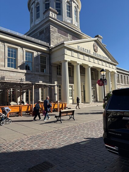 A historic building with columns and a clock tower, a paved square with benches, and people walking. 2
