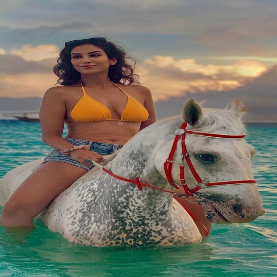 A person in a yellow bikini sitting on a white horse with red halters in turquoise water. 1
