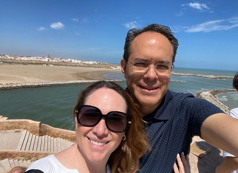A couple taking a selfie by a coastal promenade with a beach and rocky shoreline in the background. 3