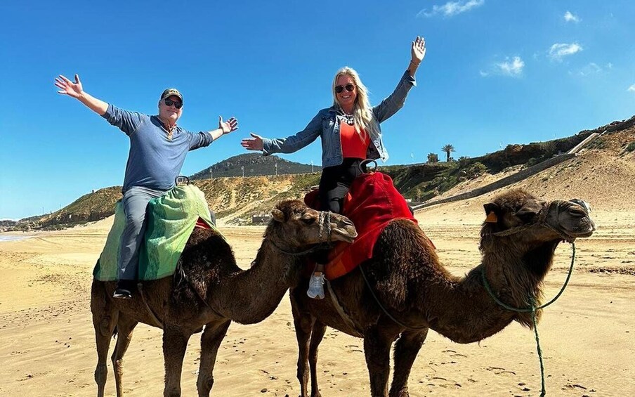 Two people riding camels on a sandy beach with a clear sky. 1