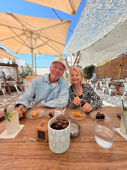 A couple dining at an outdoor café with umbrellas, a table with food and drinks, and a decorative plant. 1