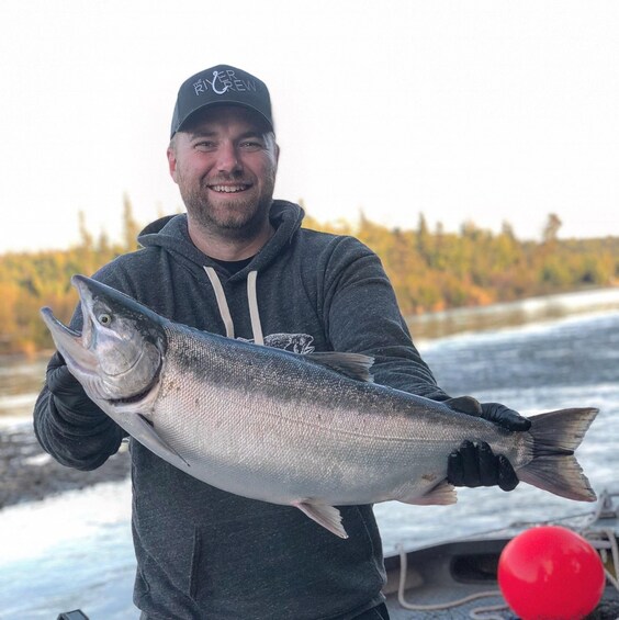 A person holding a large fish, wearing a cap with "The River Crew" logo, standing by a boat on a river with autumn trees in the background. 3