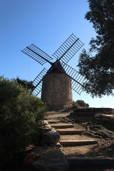 A stone windmill with wooden sails, surrounded by trees and a dirt path leading up to it. 2