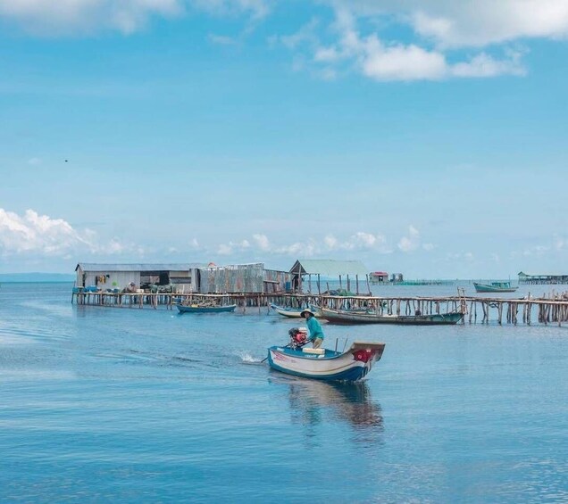 A calm sea with a small boat carrying a person. There are wooden structures and boats on the water. 5