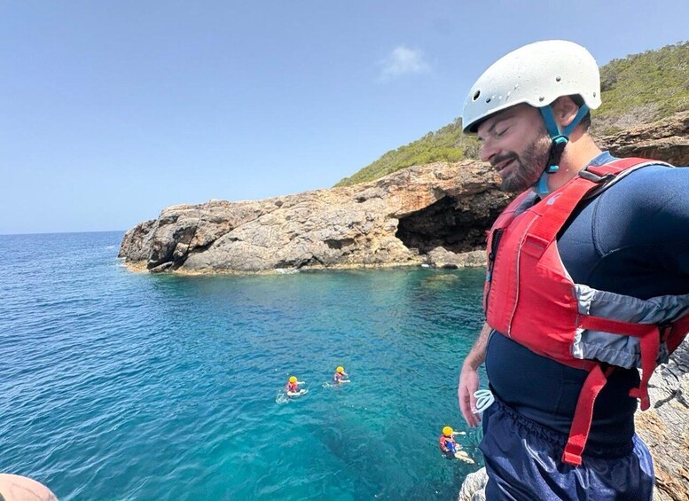 A person in a white helmet and red life jacket standing on a rocky cliff overlooking the sea, with two people in the water. 2