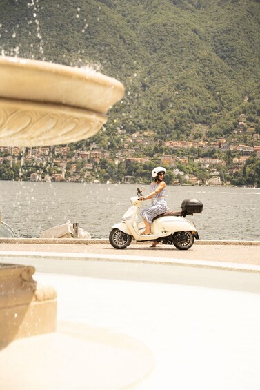 A person riding a white scooter by a body of water with a fountain in the foreground. 4