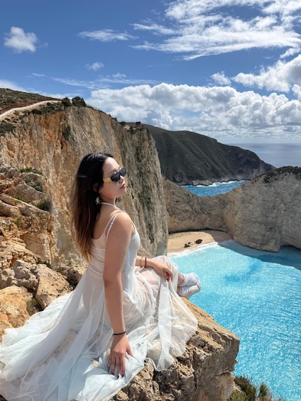 A person in a white dress sitting on a rocky cliff overlooking a clear blue sea and a rugged coastline. 2