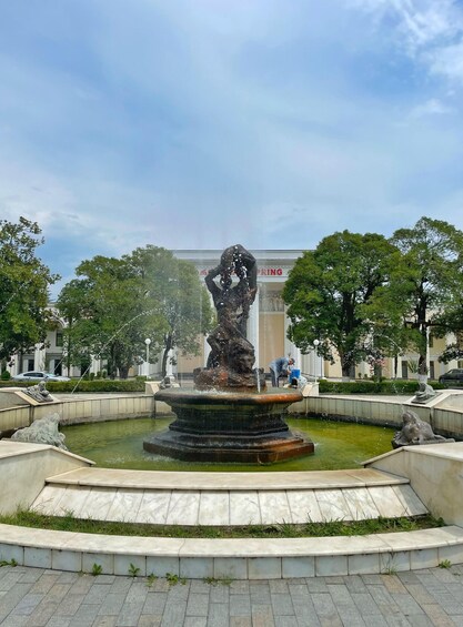 A fountain with a statue in the center, surrounded by a paved area and trees. 3