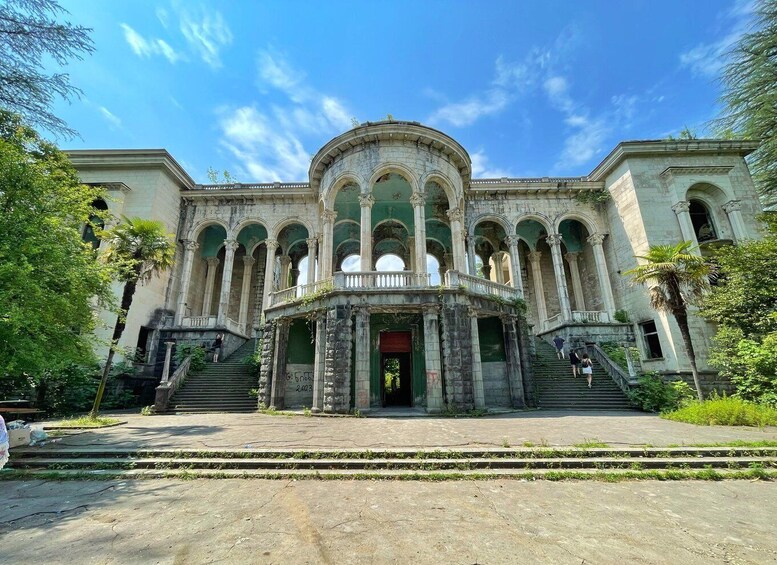 A large, historic building with multiple arches and columns, surrounded by greenery. 1