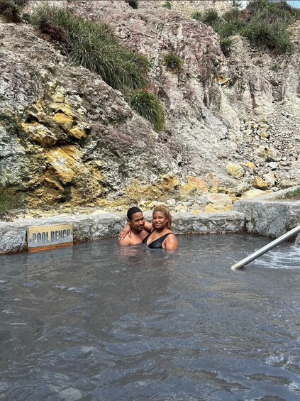 Two people in a natural hot spring with a rocky hillside in the background. 2