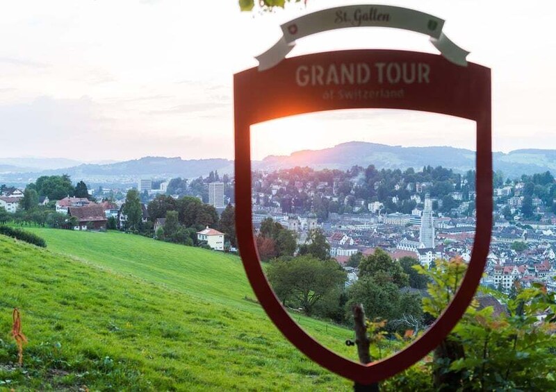 A sign for the Grand Tour of Switzerland with a scenic view of a town and hills in the background. 3