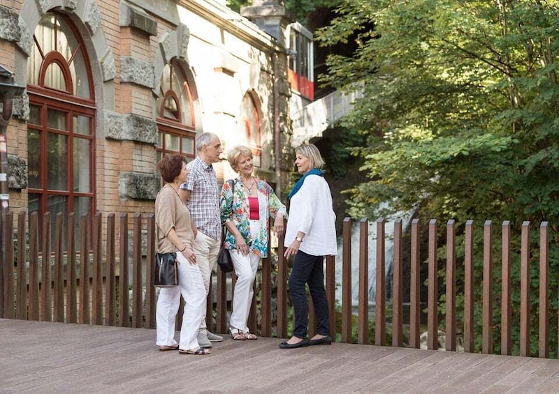 A group of people standing on a wooden deck in front of a historic building with arched windows and a brick facade. 1
