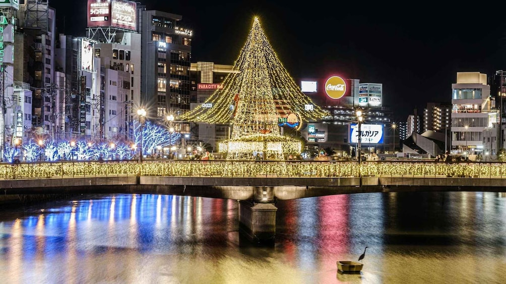 Un árbol de Navidad muy iluminado en el centro de una plaza de la ciudad, rodeado de edificios iluminados y un puente sobre una superficie acuática reflectante. 2