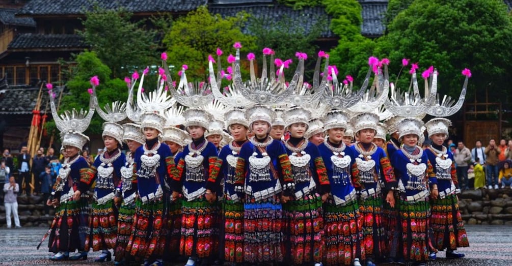 A group of people in traditional attire with elaborate headdresses and colorful skirts, standing in a line outdoors. 4