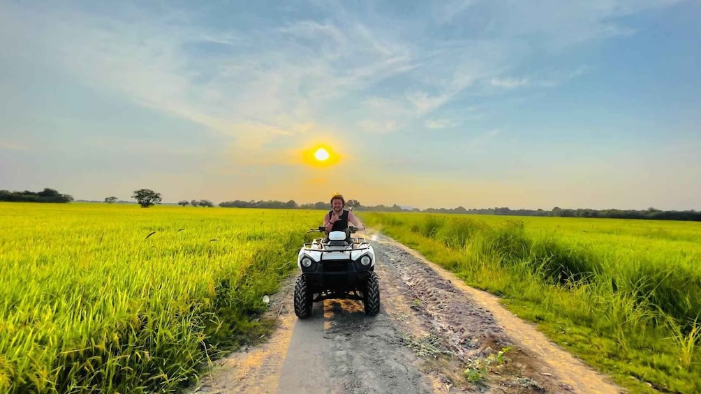 A person riding an ATV on a dirt path through a field of green crops under a clear sky. 3