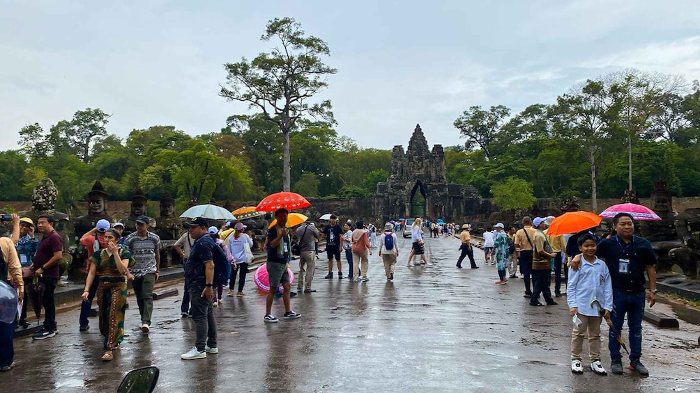 A group of people walking on a wet road with umbrellas, surrounded by trees and ancient stone structures. 5