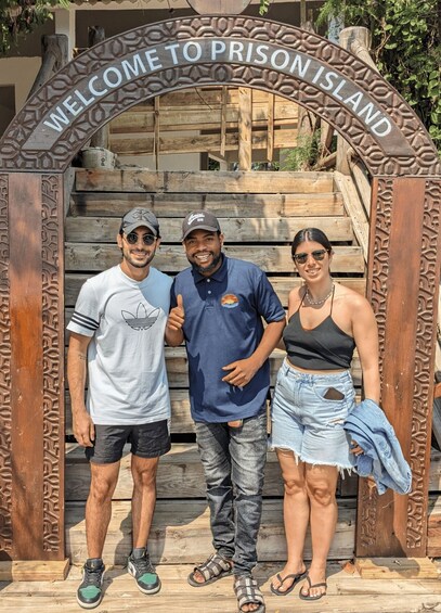 Tres personas de pie frente a un arco de madera con la inscripción "WELCOME TO PRISON ISLAND". 2
