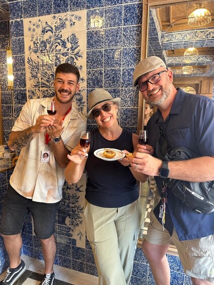 Three people holding glasses of red wine and a plate of food in a room with blue and white patterned tiles. 2