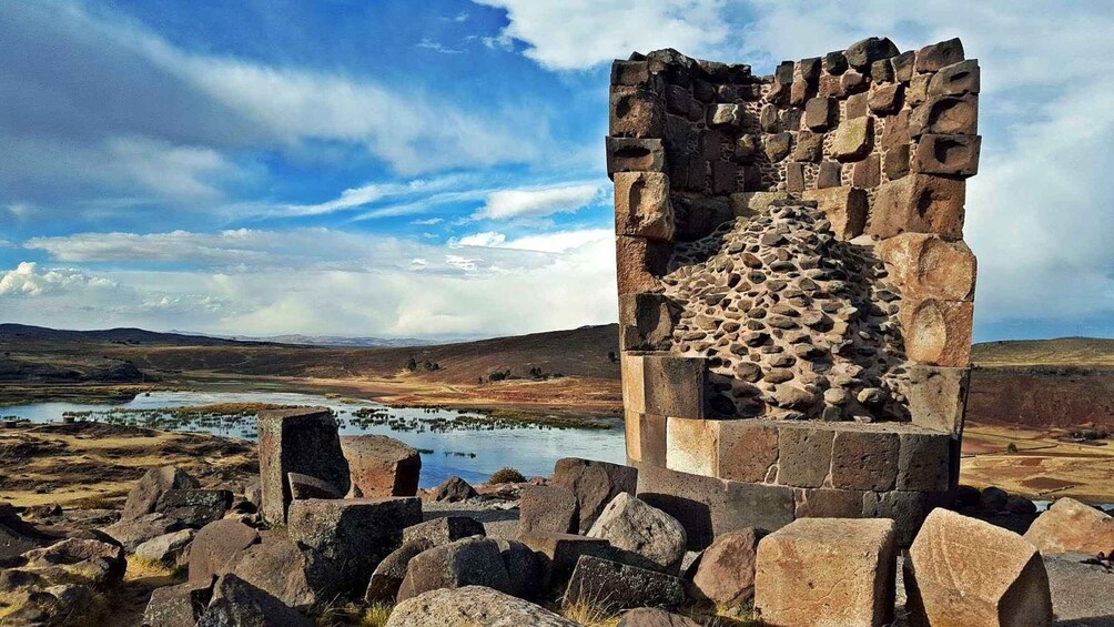 A stone ruin with a circular stone structure in the center, surrounded by rocks and a lake in a hilly landscape. 2