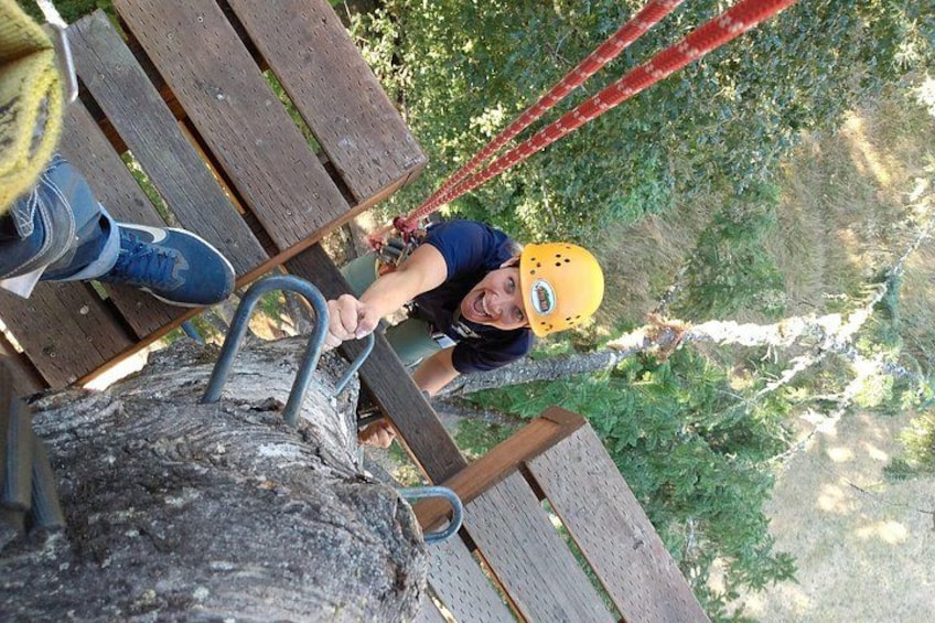 A person in a yellow helmet using a rope to climb a wooden structure in a forested area. 2