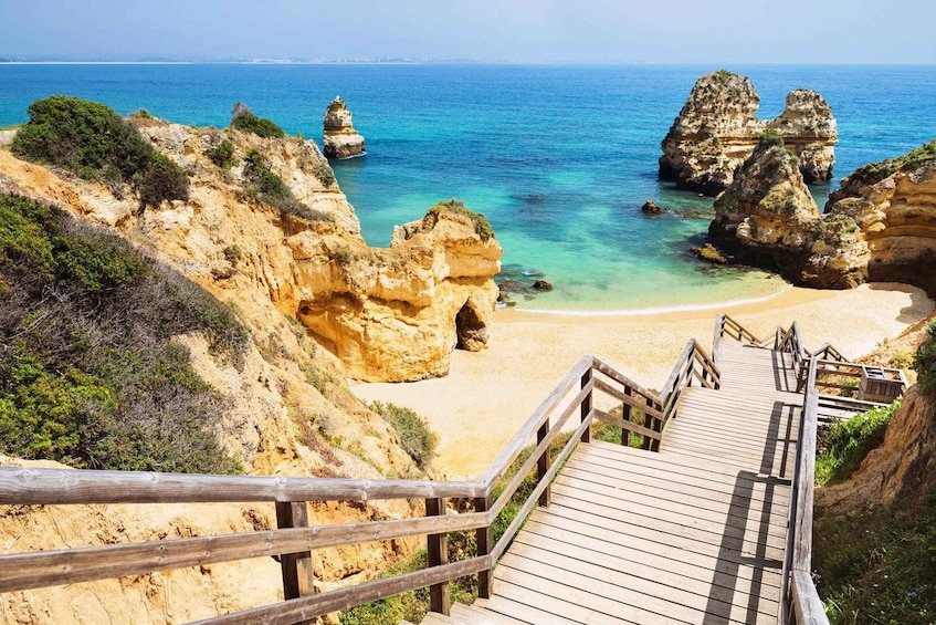 Un paysage côtier avec un escalier en bois menant à une plage de sable, entouré de falaises rocheuses et bordé d’eaux bleues et limpides. 1